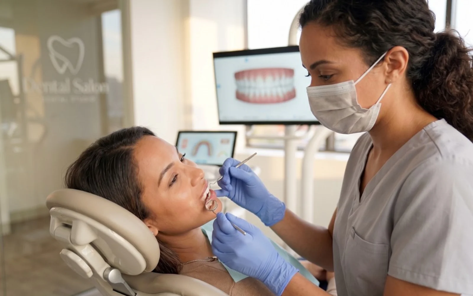 Dental professional examining a patient's teeth with a mirror and probe, with a 3D dental scan on a monitor in the background.
